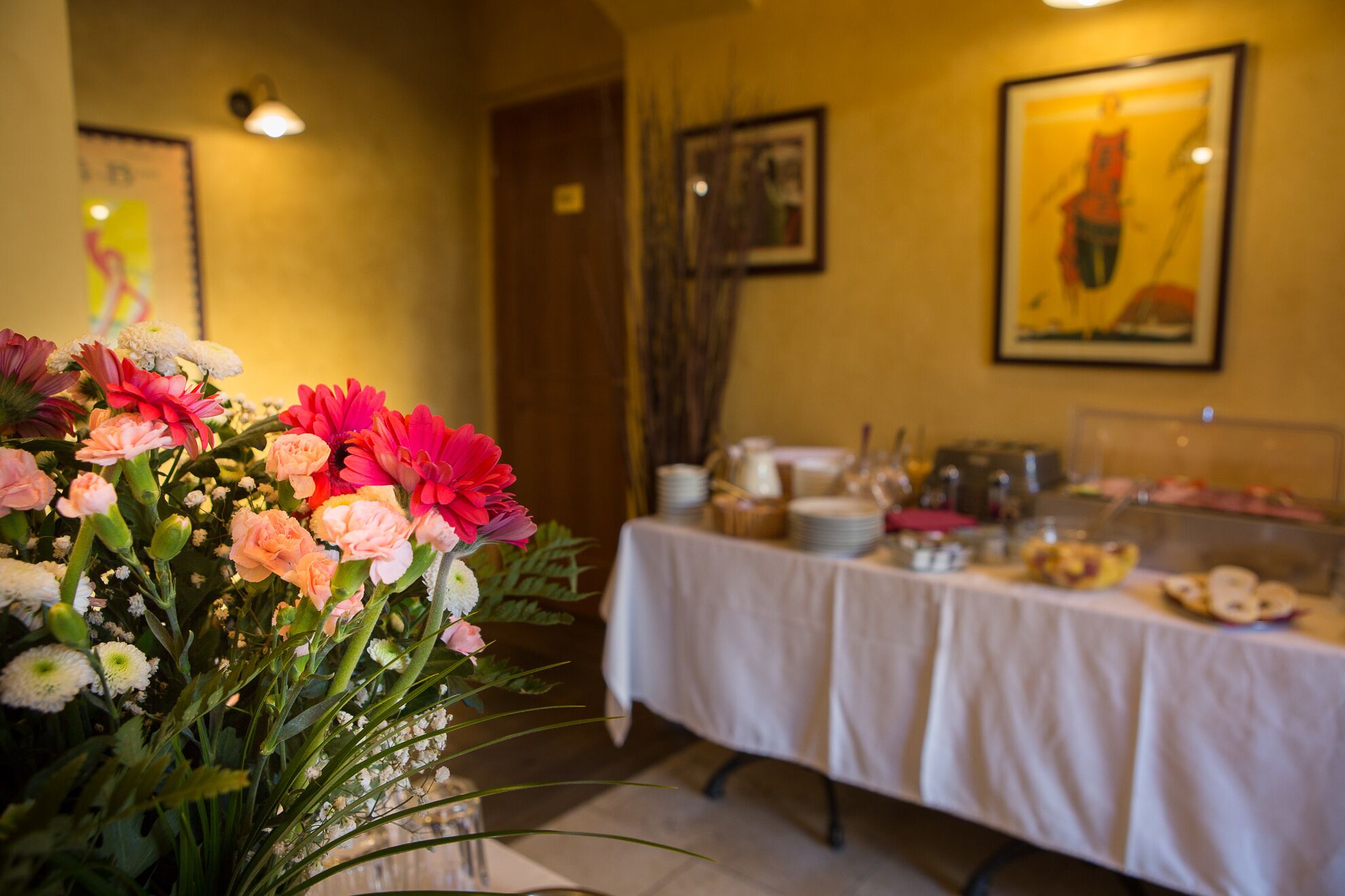 A dining area with a table set for a meal, a floral arrangement in the foreground, and artwork on the walls.