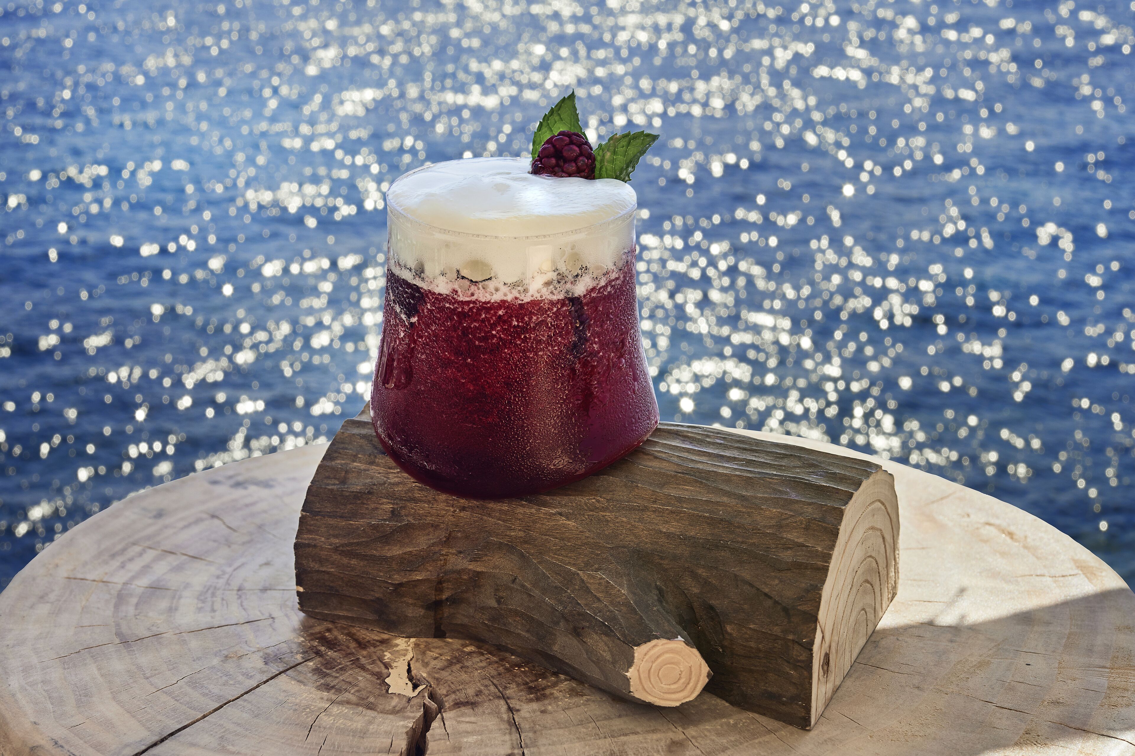 Un verre d’une boisson rouge, agrémenté de quelques baies, posé sur une surface en bois, à proximité d’une étendue d’eau scintillante.
