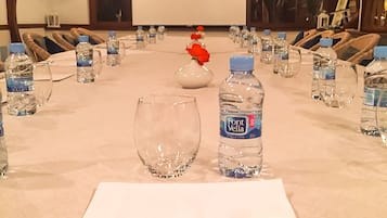 Un salón de conferencias con una larga mesa, botellas de agua, un bolígrafo y un jarrón con flores rojas.