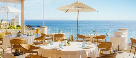 An outdoor dining area with round tables and wicker chairs, overlooking the sea.
