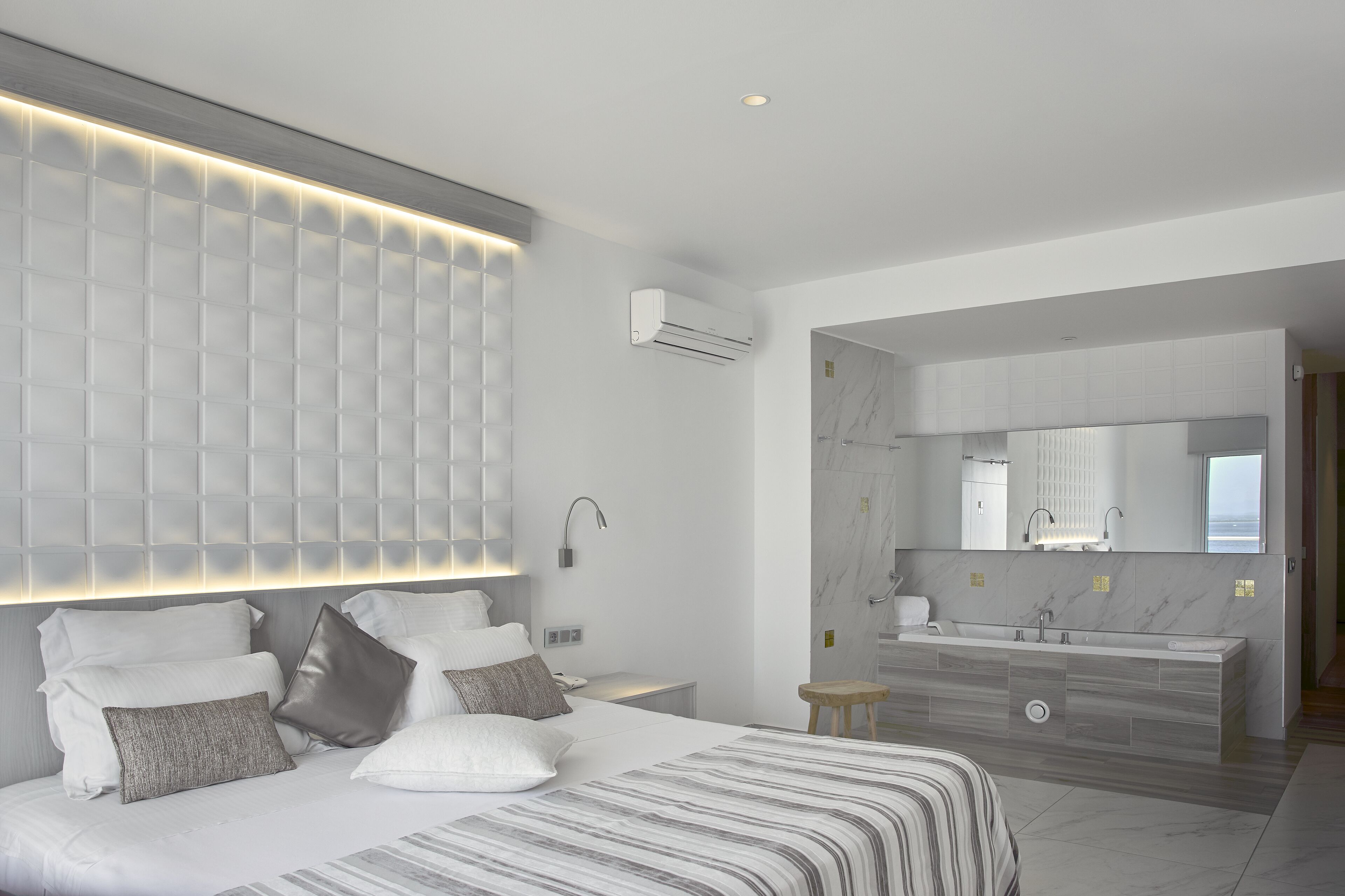 Una habitación de hotel moderna con una cama grande, un cabecero integrado, un baño con un espejo amplio y suelo de mármol.