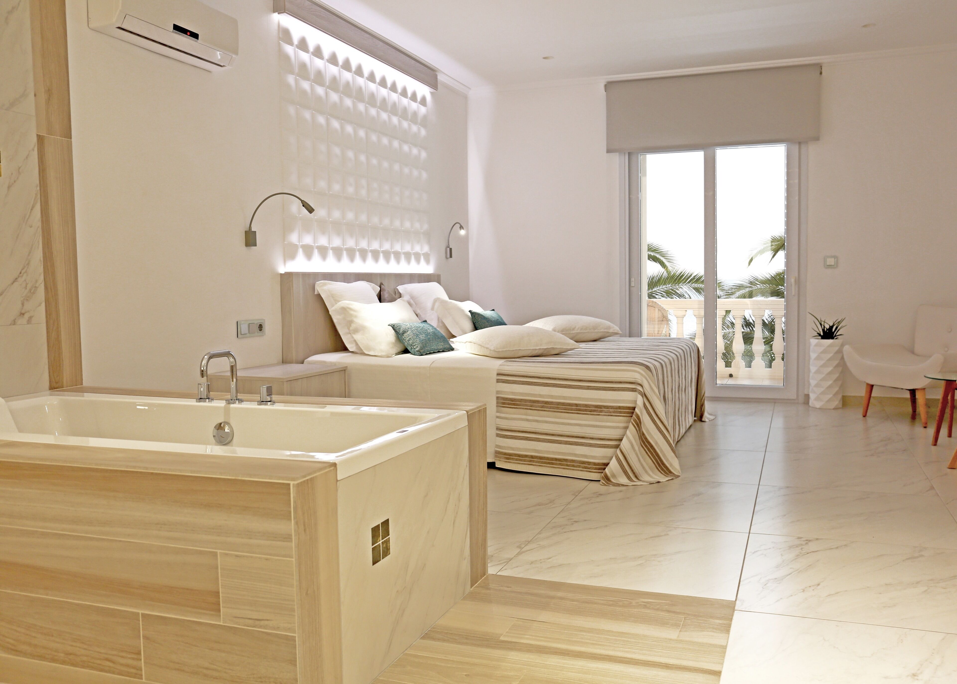 A modern bedroom with a large bathtub, a bed with multiple pillows, and a balcony with a view.