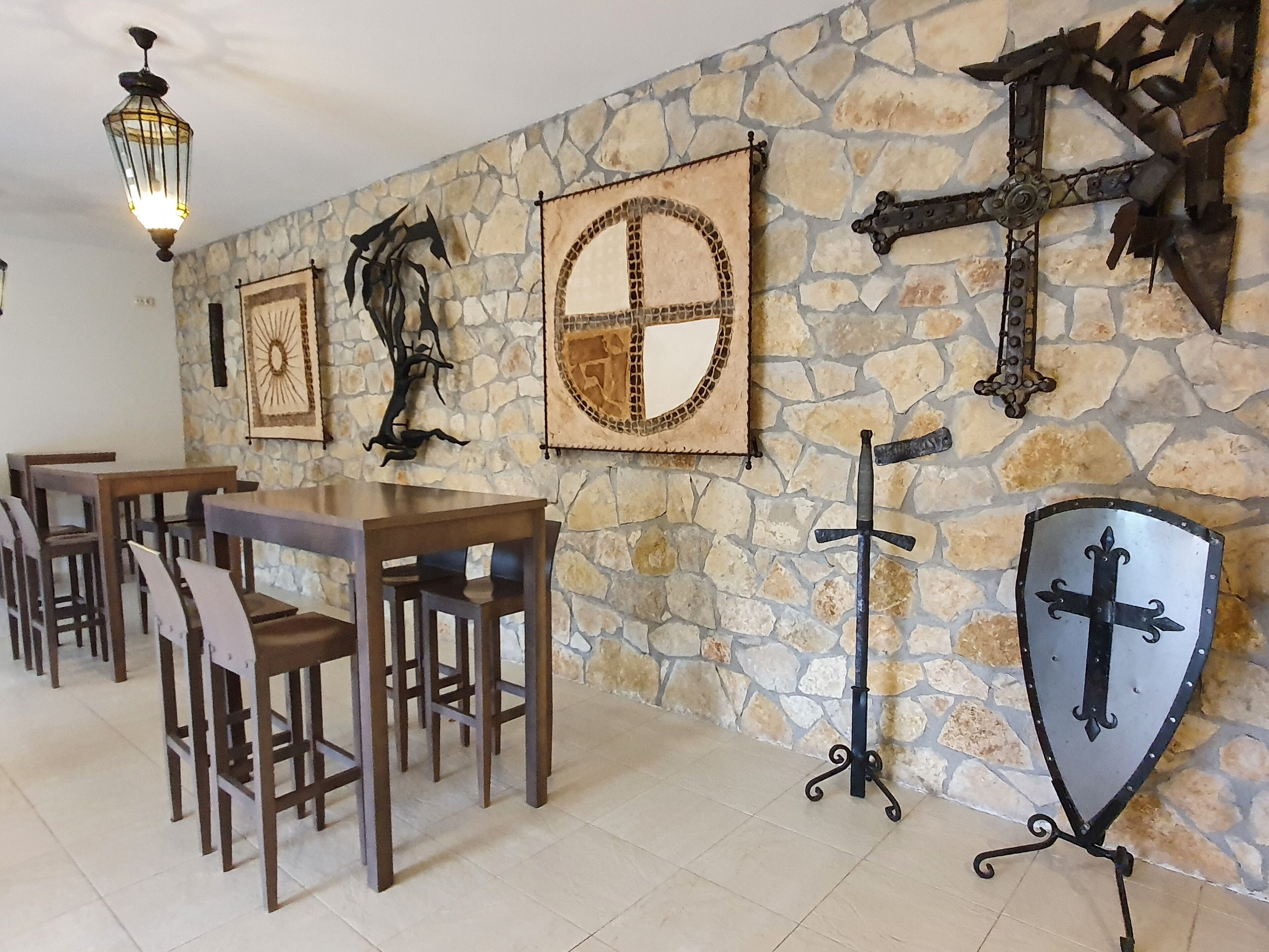 A stone wall with medieval-themed wall art, including shields and swords. There are wooden tables and chairs arranged for dining.