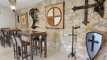 A stone wall with medieval-themed wall art, including shields and swords. There are wooden tables and chairs arranged for dining.