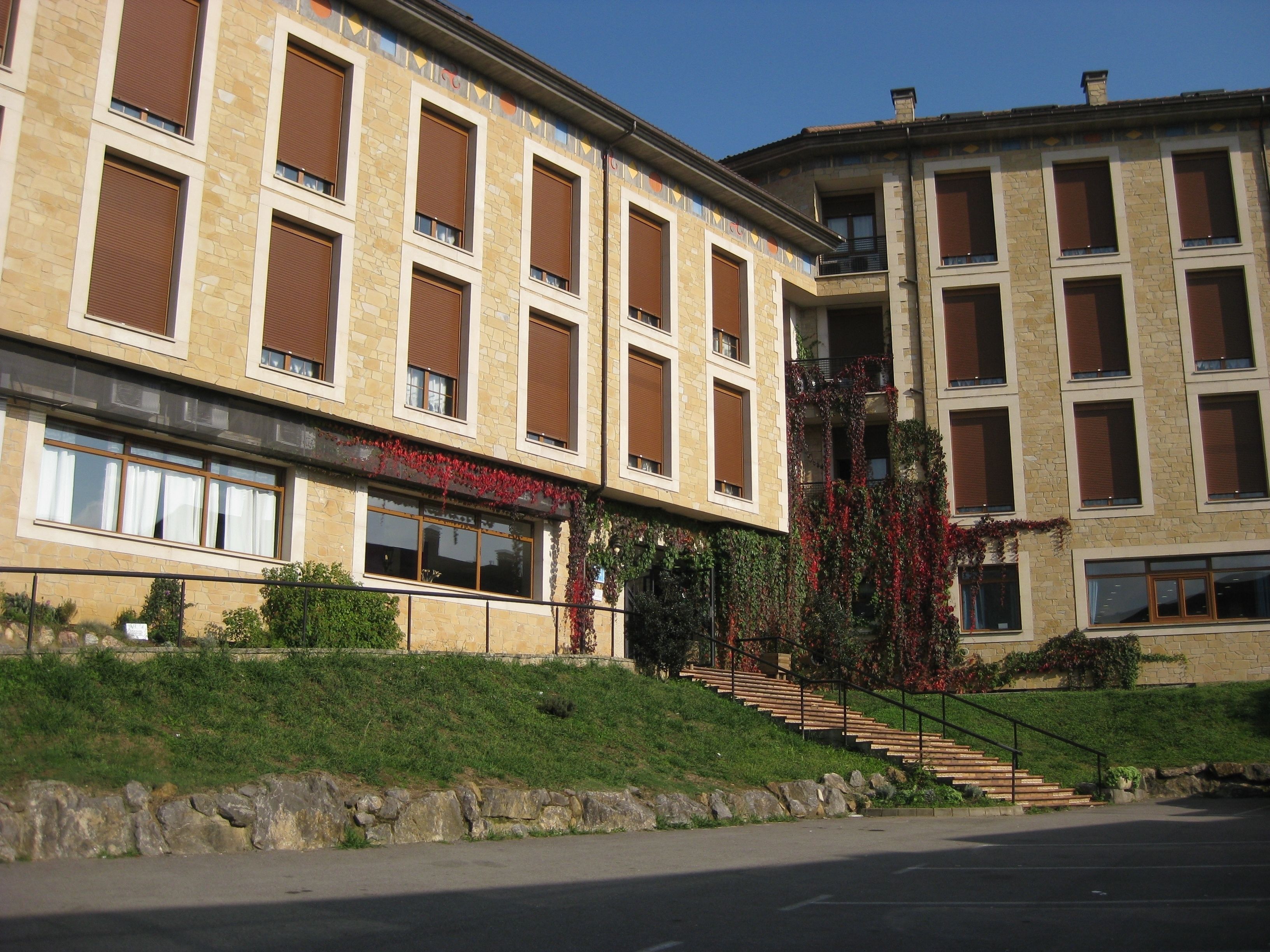 Un edificio a più piani con facciata in pietra, ampie finestre dotate di persiane marroni e una scala che conduce a un balcone.