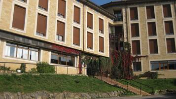 Un edificio a più piani con facciata in pietra, ampie finestre dotate di persiane marroni e una scala che conduce a un balcone.