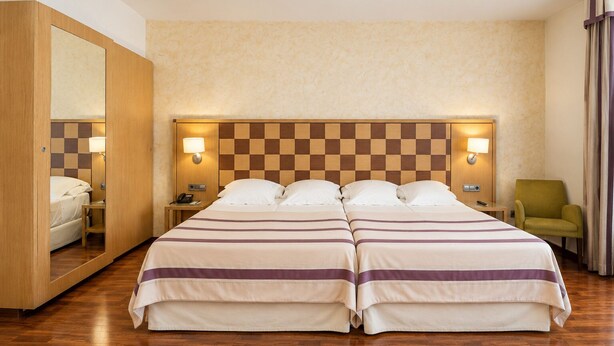 A hotel room with two beds, wooden headboard, bedside lamps, a mirror, a chair, and a wooden floor. - Duran Hotel & Restaurant (Figueres)