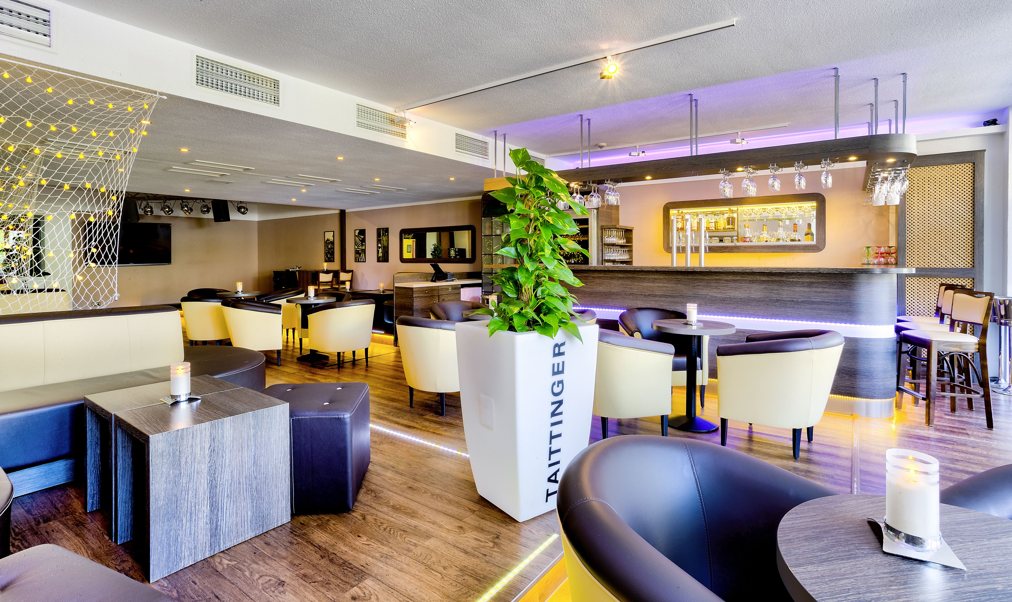 Un hall d’hôtel moderne, doté d’espaces aménagés avec des sièges, d’un bar et d’un grand bac à plantes contenant une plante.