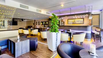 Un hall d’hôtel moderne, doté d’espaces aménagés avec des sièges, d’un bar et d’un grand bac à plantes contenant une plante.