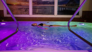 Una persona recostada en una piscina con vista a la puesta de sol a través de una ventana.