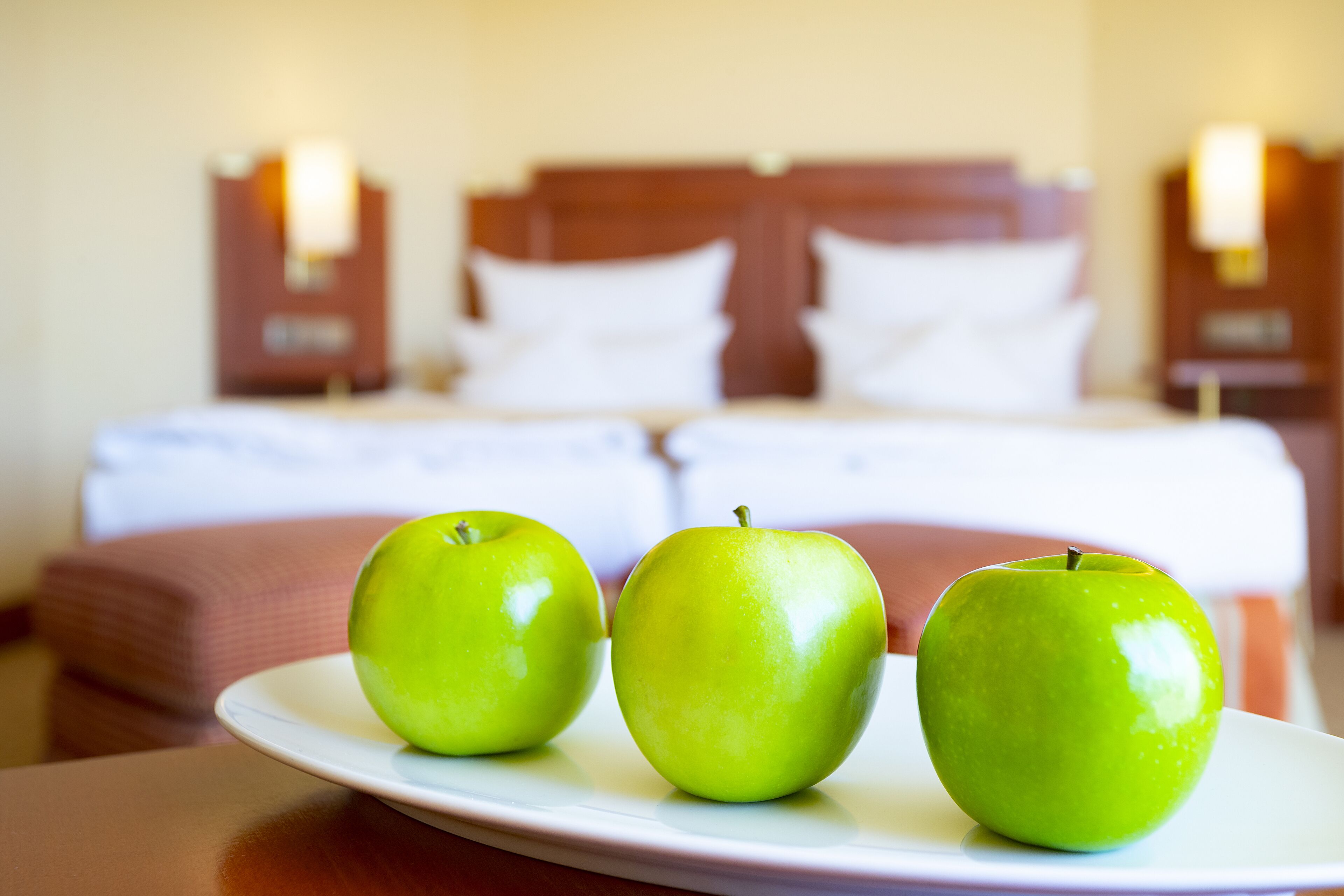 A hotel room with a bed, pillows, and a plate with three green apples on a table.