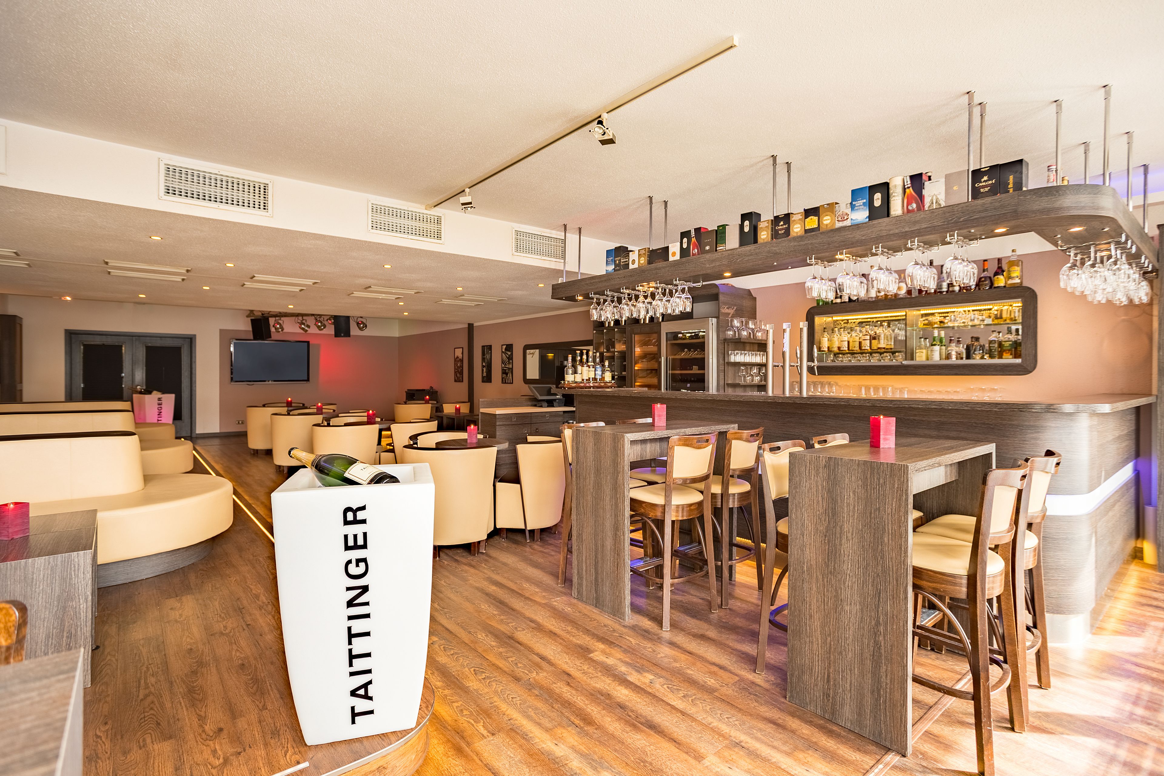 A modern bar area with a wooden floor, bar stools, and a variety of bottles and glasses on shelves.