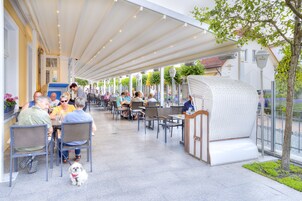 An outdoor café with tables and chairs under a covered area, surrounded by greenery and a building with a sign.