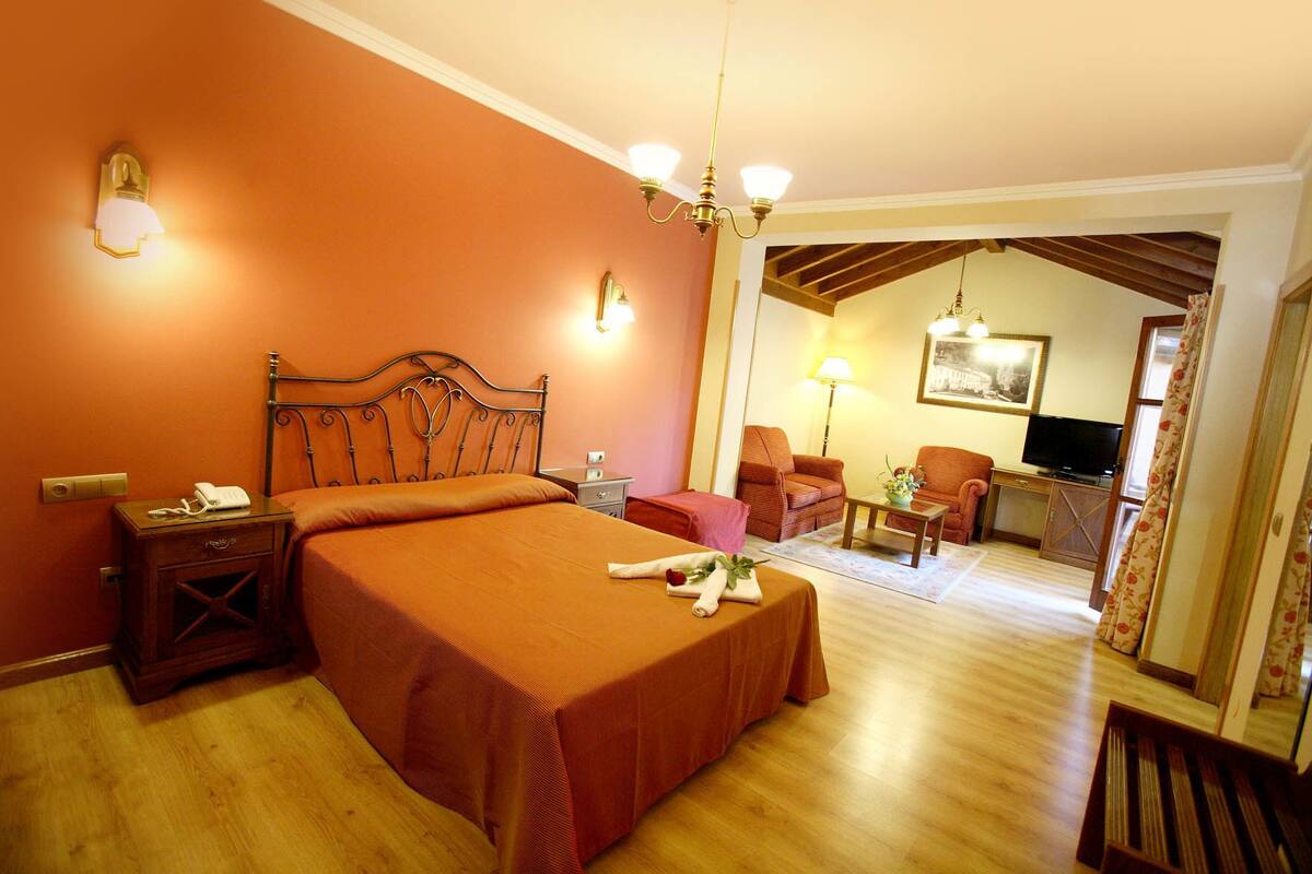Una habitación de hotel con una cama grande, una mesita de noche de madera, un escritorio con lámpara, dos sillones, un televisor y un cuadro en la pared.