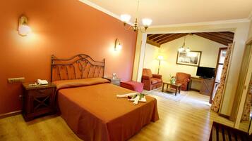 Una habitación de hotel con una cama grande, una mesita de noche de madera, un escritorio con lámpara, dos sillones, un televisor y un cuadro en la pared.
