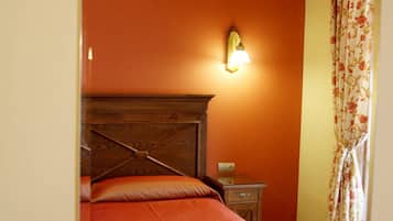 A bedroom with a wooden headboard, a single bed with a red bedspread, a nightstand, a wall-mounted lamp, and a floral curtain.