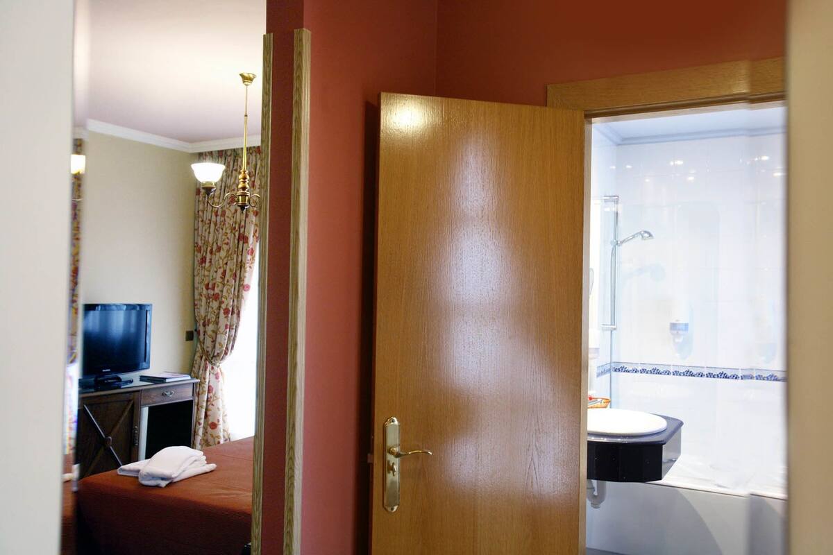 Habitación de hotel con puerta de madera, televisor sobre un escritorio, baño con lavamanos y espejo, y ducha.