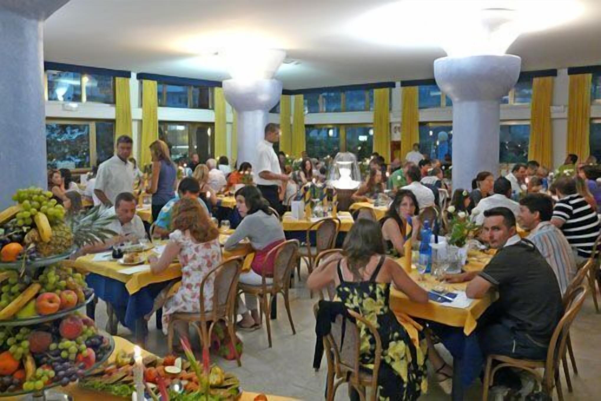 A bustling restaurant with multiple tables and chairs, a fruit display, and large windows in the background.