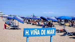 Une plage avec de nombreux parasols colorés et des chaises longues, une pancarte indiquant « H. VINA DE MAR » et une statue en arrière-plan.
