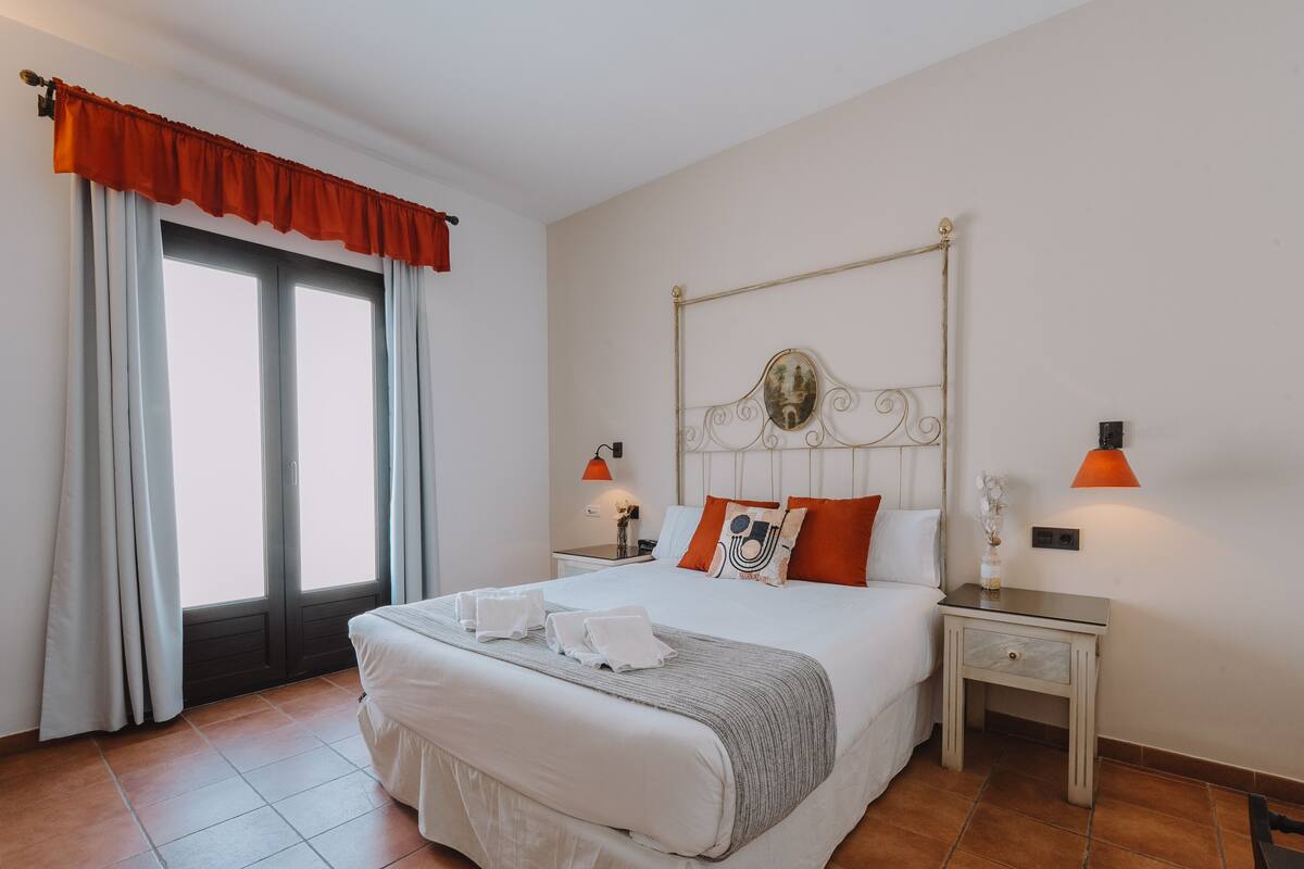 A hotel room with a large bed, two bedside tables, a nightstand, a window with curtains, and a wall-mounted lamp.