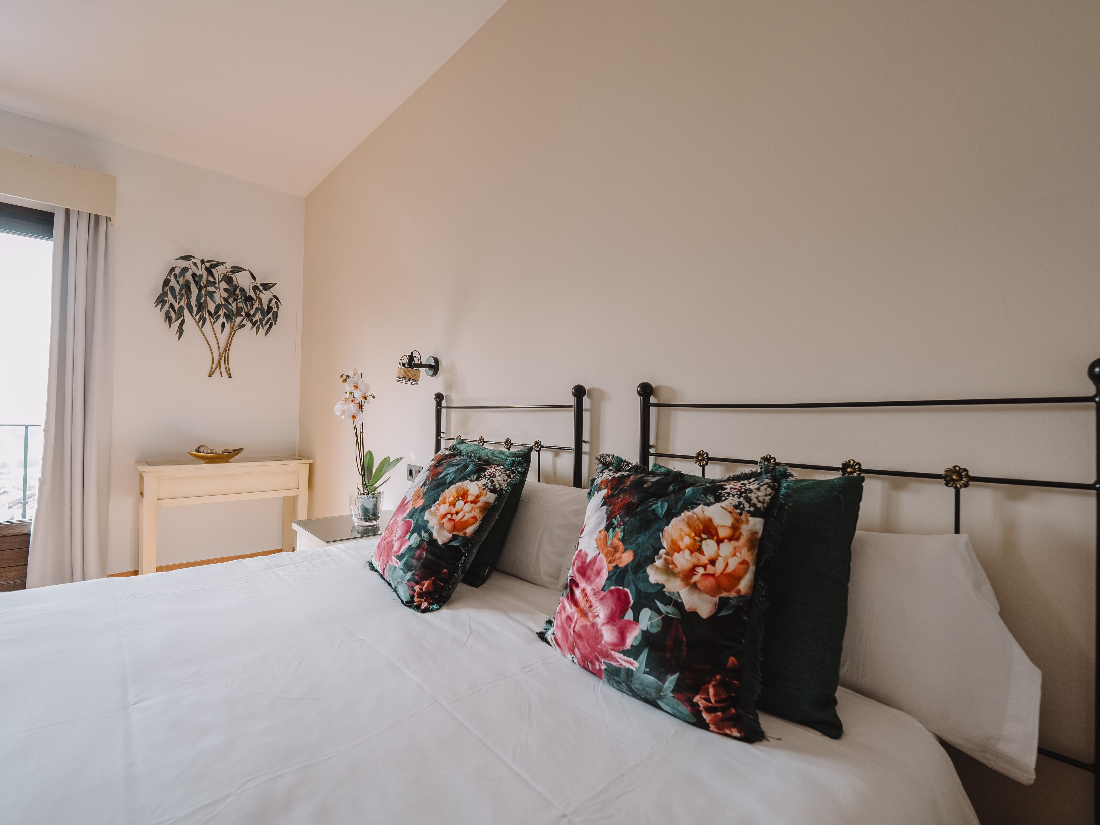 Una cama bien hecha con almohadones florales, una mesita de noche con un cuenco y una planta colgante en la pared.
