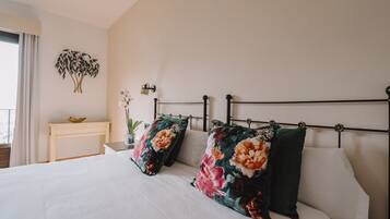 Una cama bien hecha con almohadones florales, una mesita de noche con un cuenco y una planta colgante en la pared.