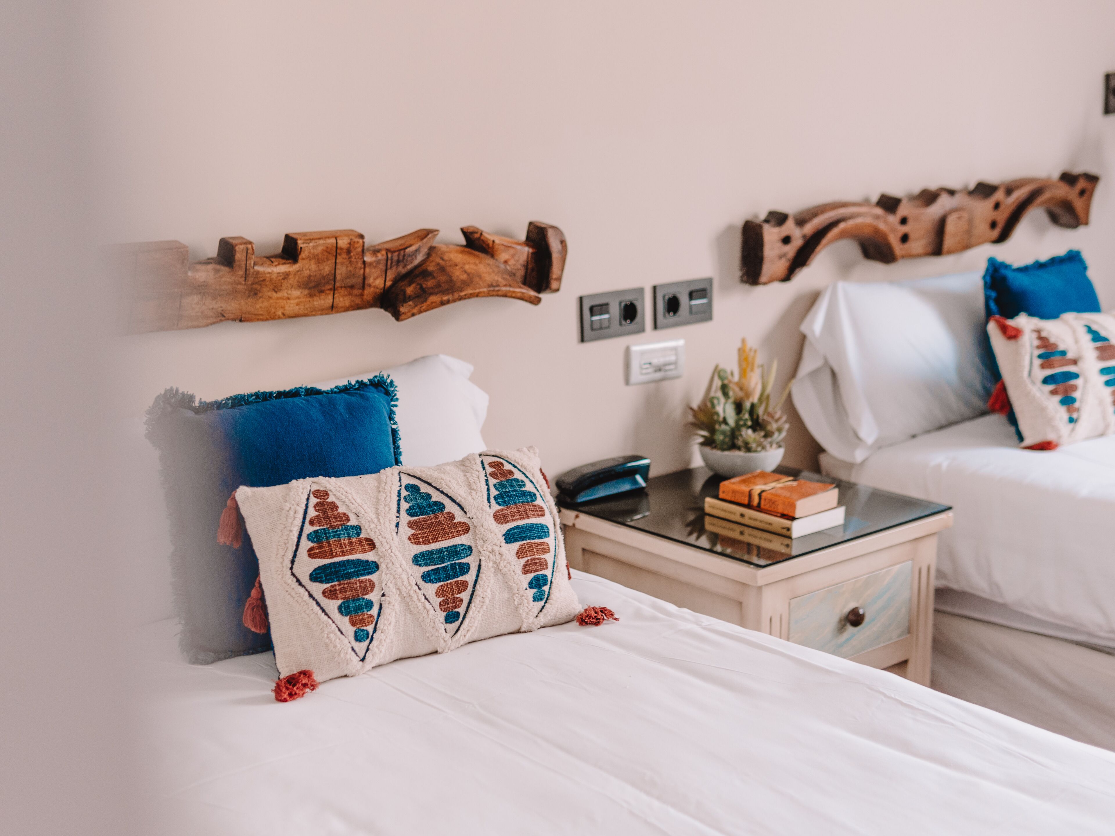Habitación de hotel con dos camas, cada una con ropa de cama blanca y almohadas decorativas. Se observa un cabecero de madera a la izquierda, una pequeña mesa con una lámpara, y una repisa con libros y una planta.