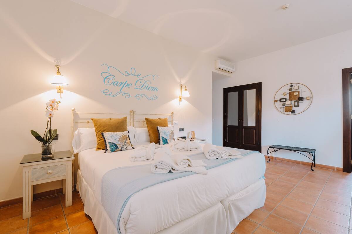A hotel room with a bed, bedside tables, a wall-mounted air conditioner, a bench, and a decorative wall piece.
