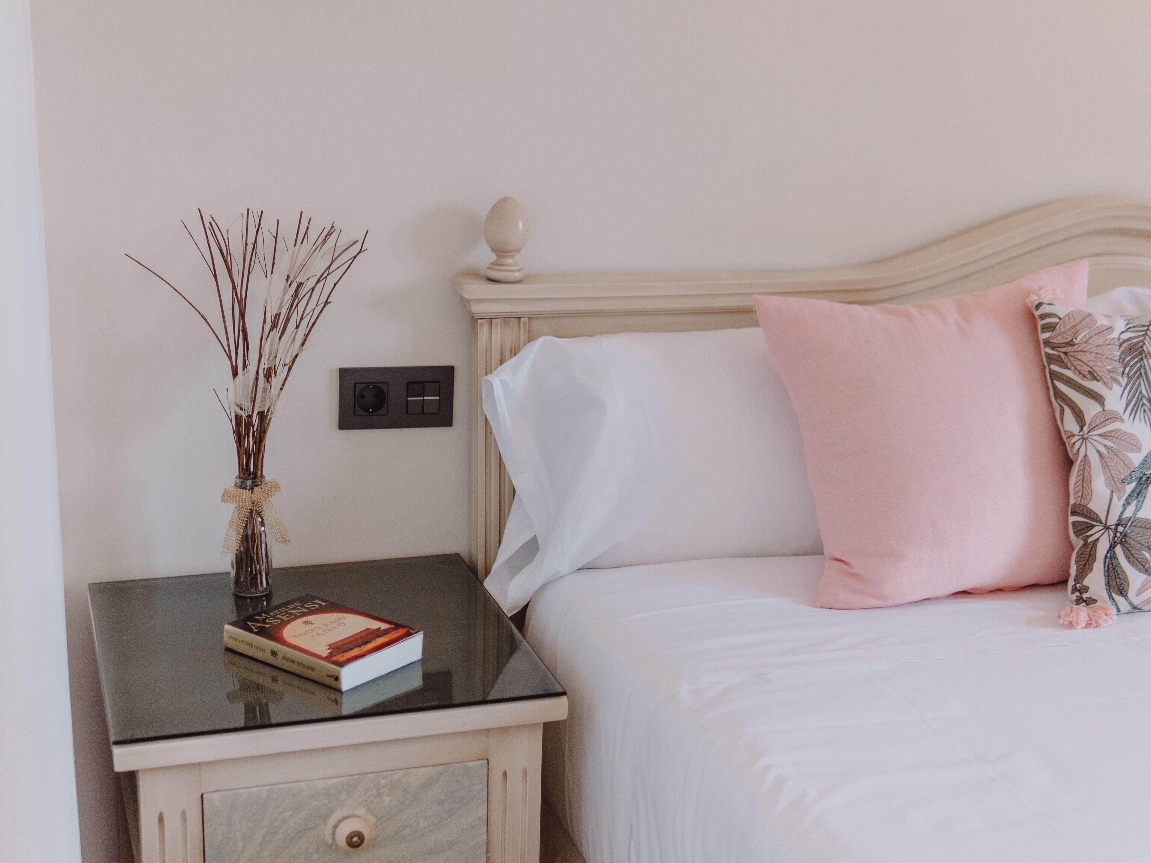 Una cama bien hecha con almohadas blancas y rosas, una mesita de noche con un libro y un jarrón con ramas secas, e interruptor de luz en la pared.