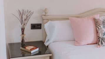 Una cama bien hecha con almohadas blancas y rosas, una mesita de noche con un libro y un jarrón con ramas secas, e interruptor de luz en la pared.