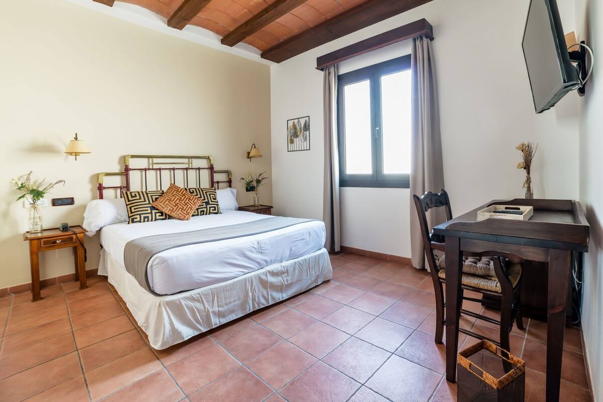 A bedroom with a bed, bedside tables, a desk, a chair, a window with curtains, and a wall-mounted TV.
