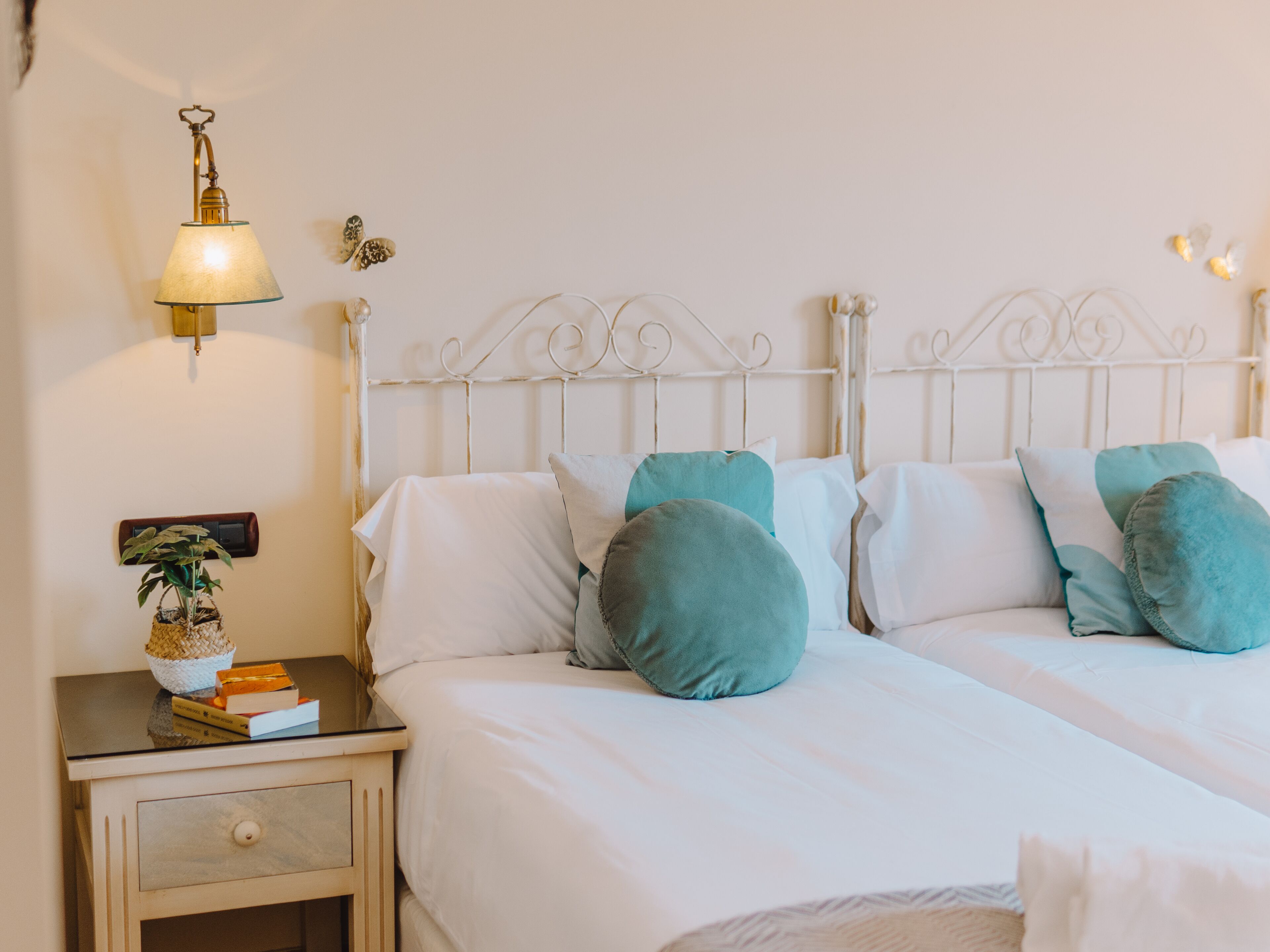 A neatly made twin bed with white bedding and teal pillows, a bedside table with a lamp, a small plant, and a few books.