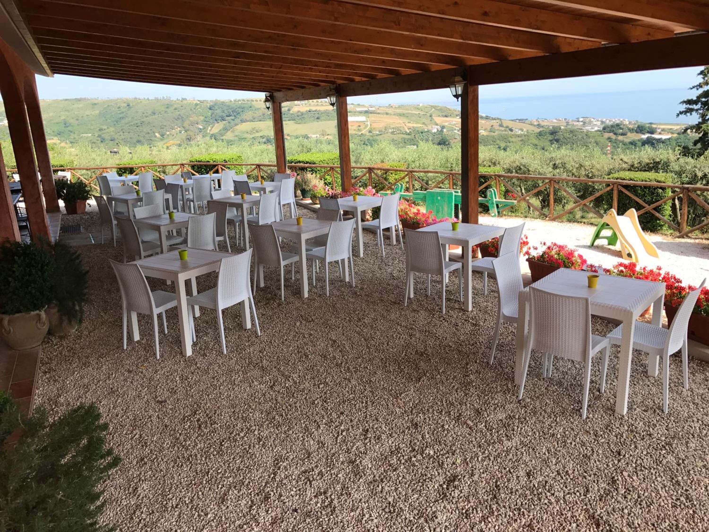 Un patio al aire libre con mesas y sillas blancas, suelo de grava y vistas a colinas verdes y al mar.