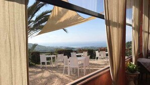 A room with a large window overlooking a patio with white chairs and tables, a view of the landscape, and a bed with a plant beside it. - I Cardinali (Santa Marinella)