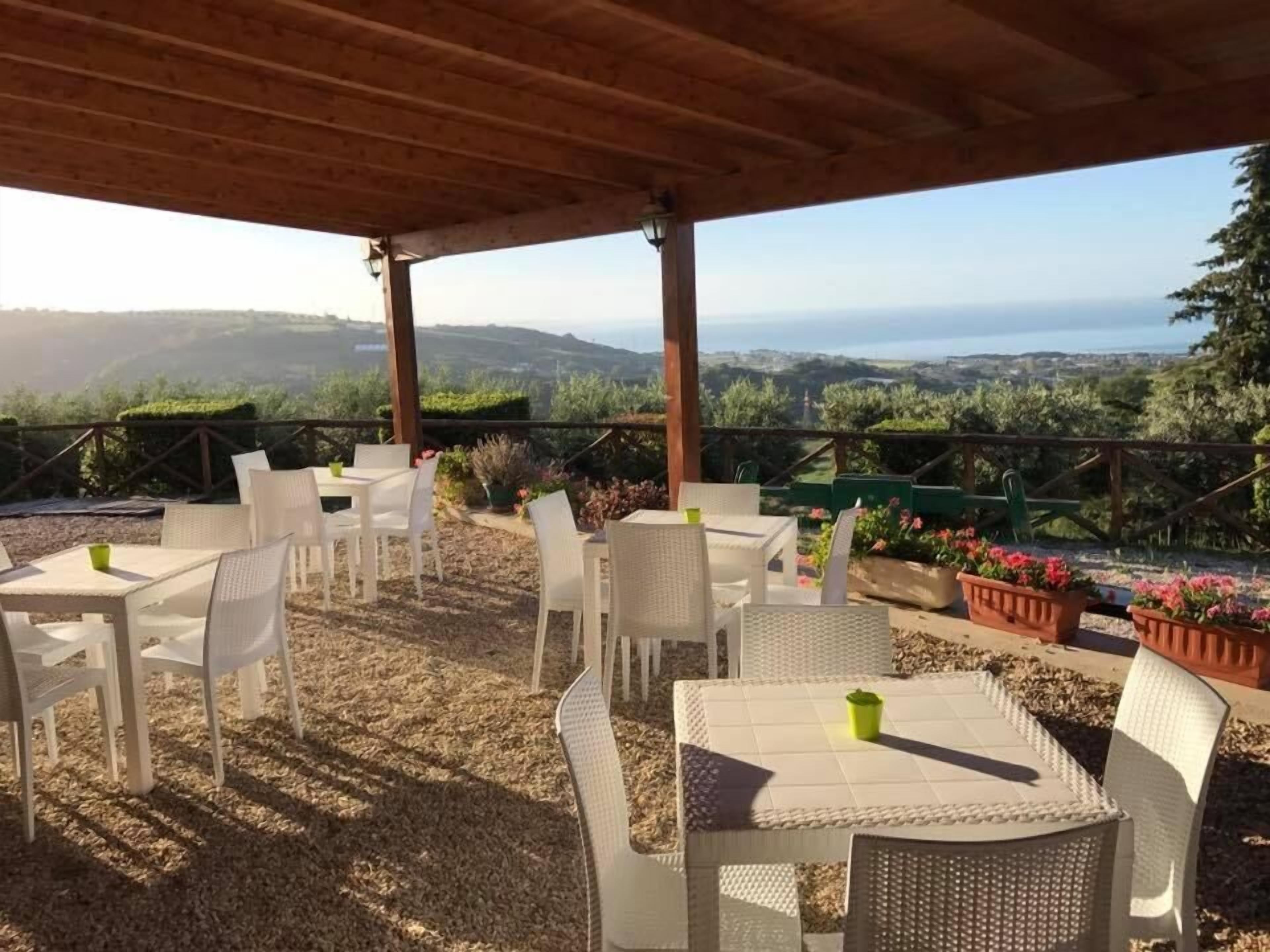 Un patio exterior con mobiliario y mesas de mimbre blanco, con vistas a un paisaje pintoresco con colinas y el mar.