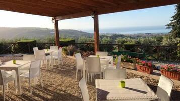 Un patio exterior con mobiliario y mesas de mimbre blanco, con vistas a un paisaje pintoresco con colinas y el mar.