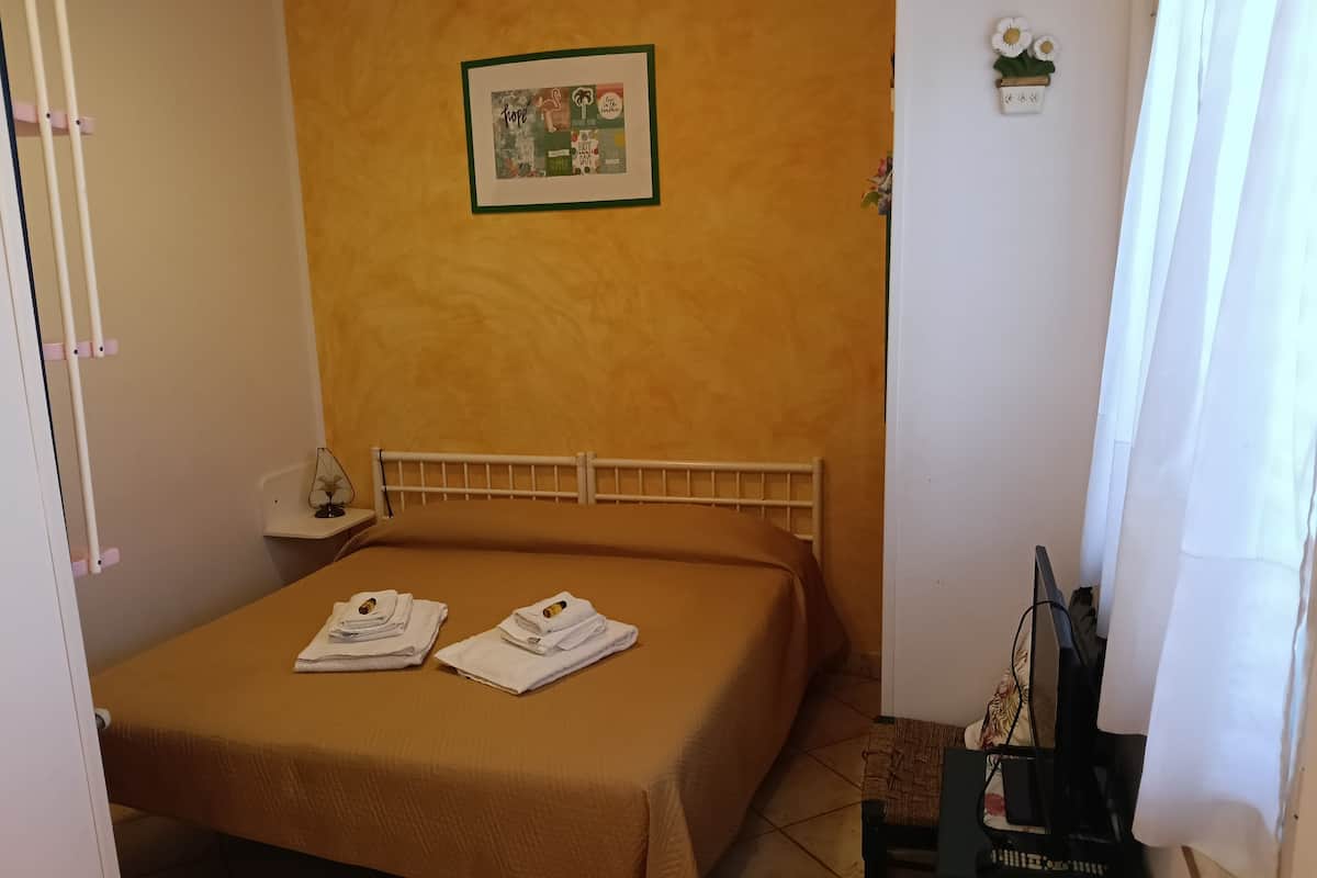 Ein kleines, ordentlich gemachtes Bett mit einer braunen Tagesdecke, zwei gefaltete Handtücher, ein gerahmtes Bild an der Wand und eine kleine Pflanze auf dem Nachttisch.