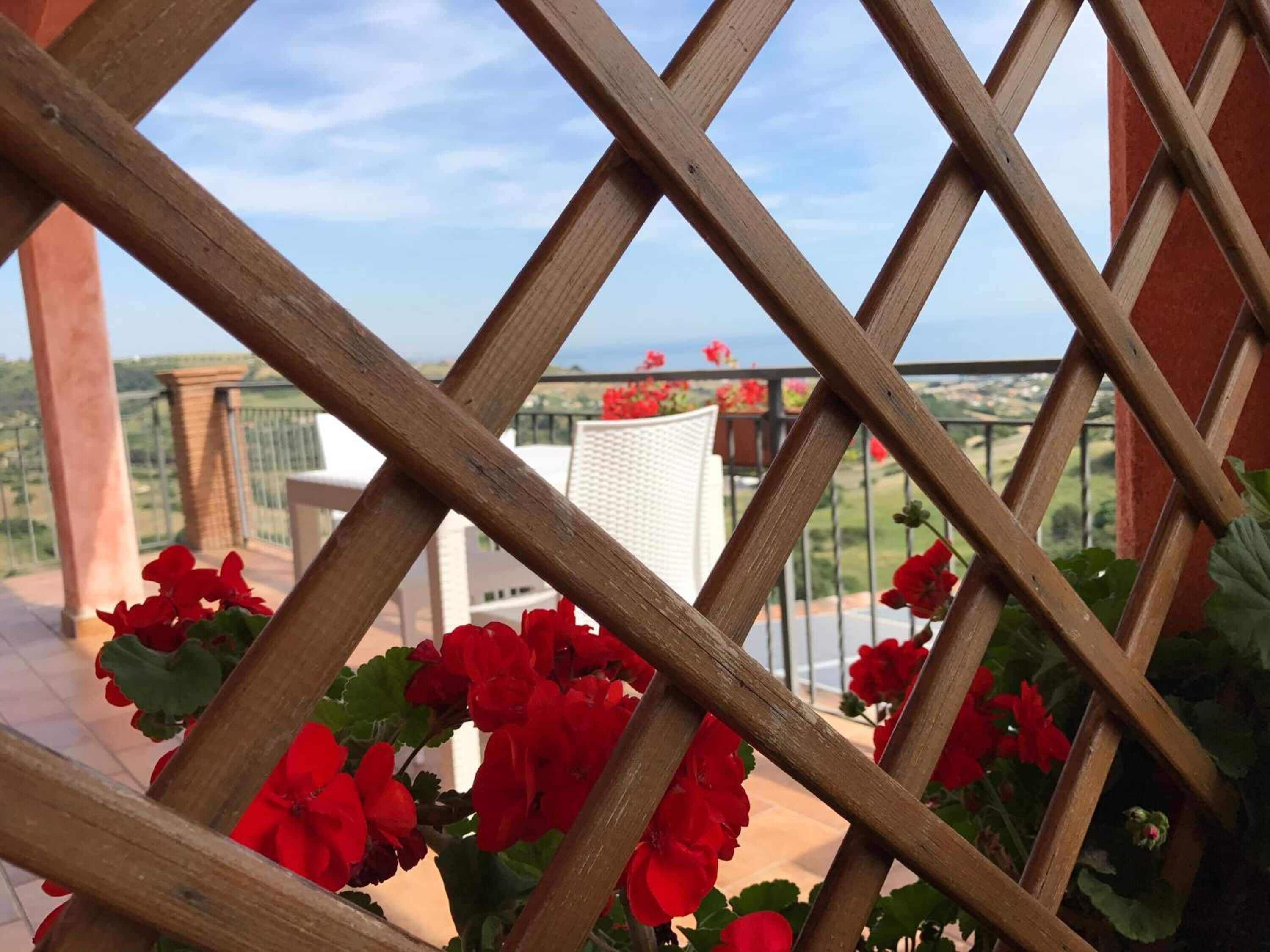 Una estructura de celosía de madera con flores rojas en primer plano, una silla blanca y, al fondo, una maceta roja y follaje verde.