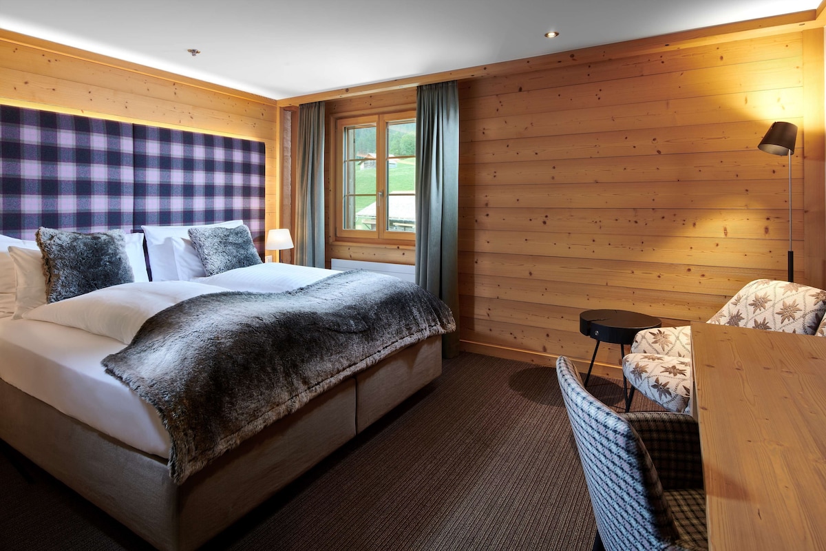 Una habitación con una cama grande, cabecera de madera, una mesita pequeña, una silla y un ventanal con cortinas.