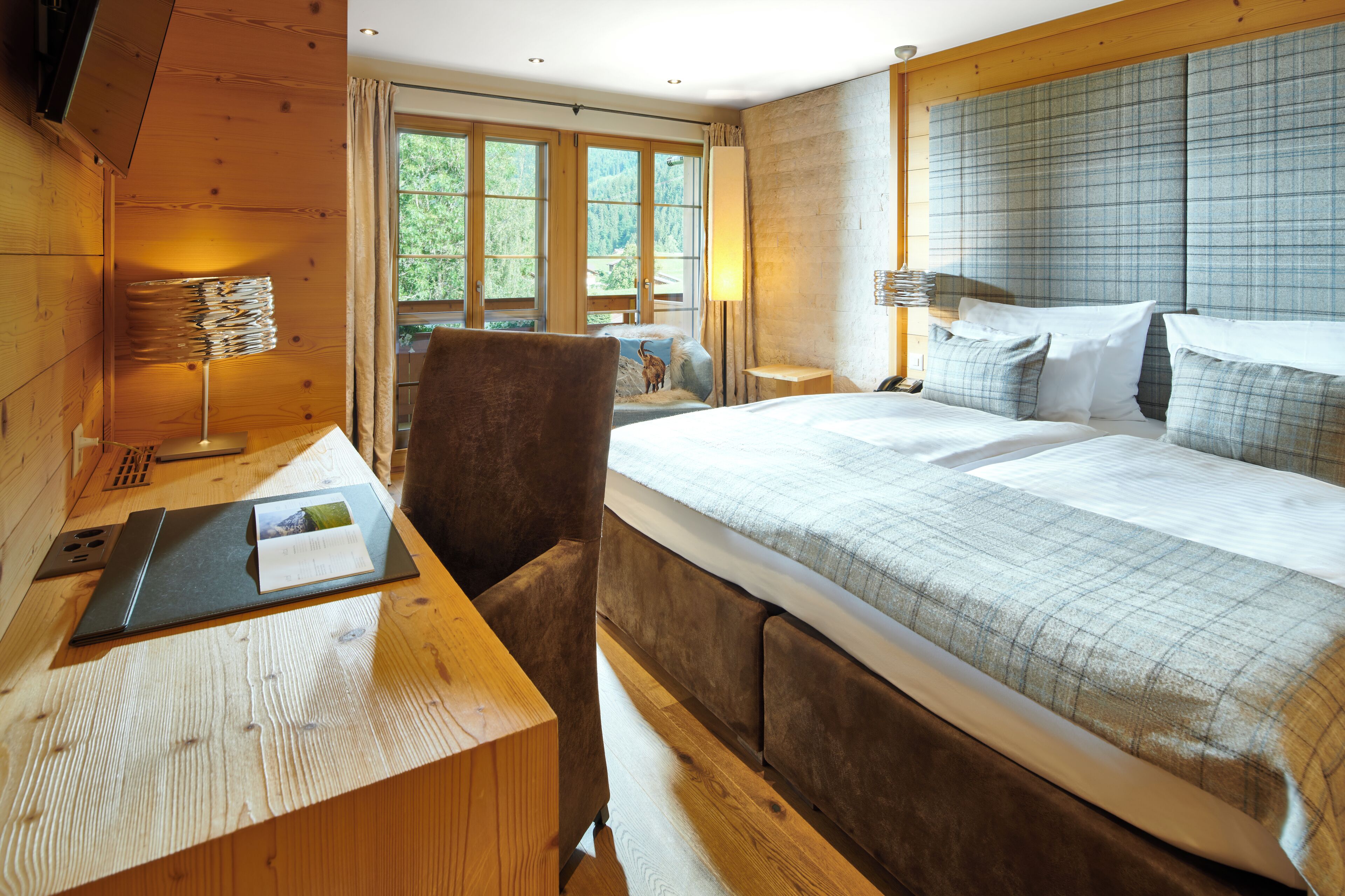 A bedroom with a large bed, wooden walls, a desk with a lamp, and a window with a view of trees.