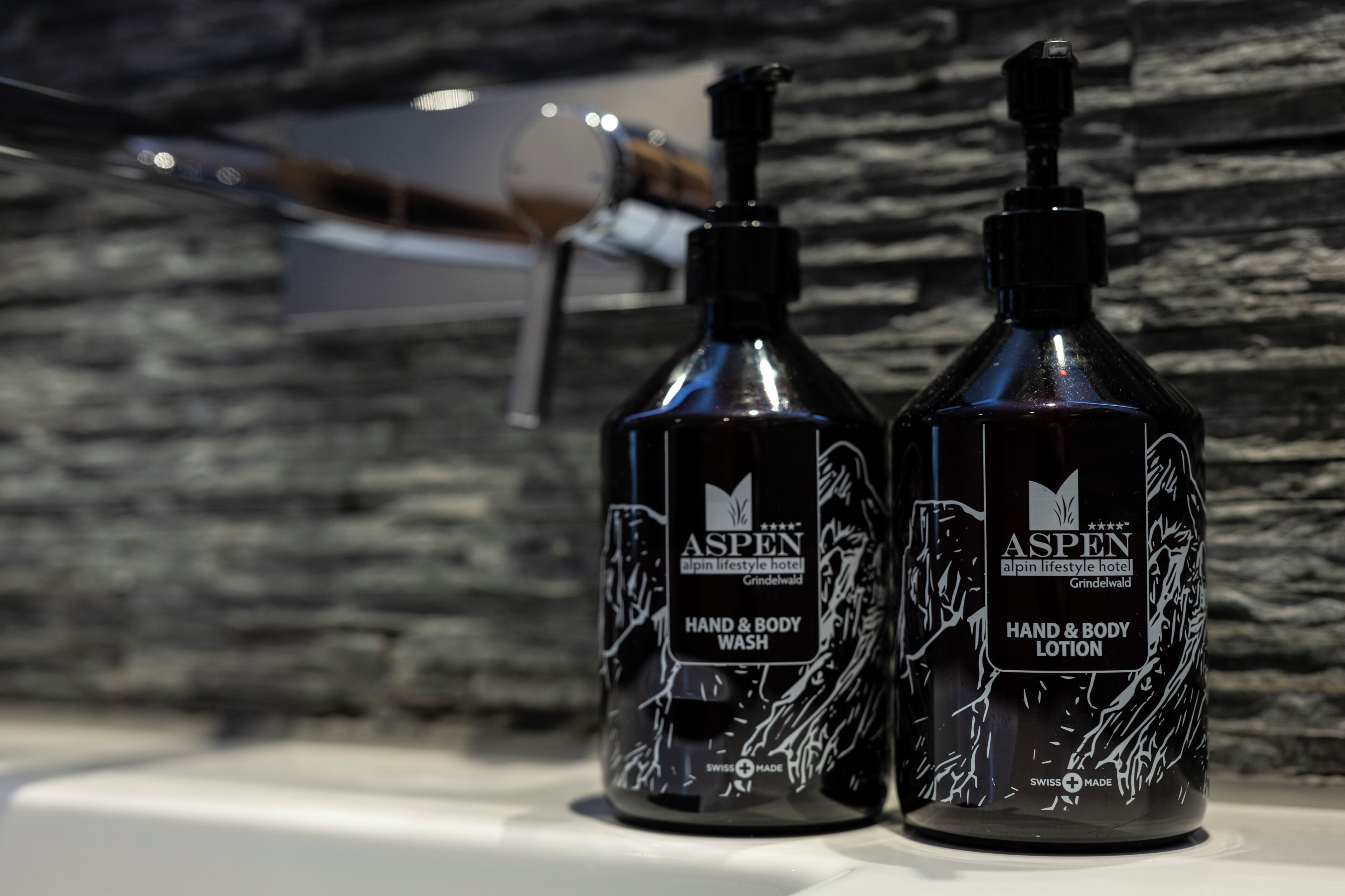 Two bottles of Aspen hand and body wash on a white surface with a stone wall background.