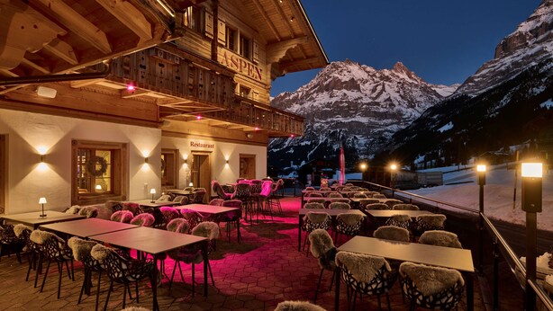 A restaurant with outdoor seating, wooden architecture, and a view of snow-covered mountains.