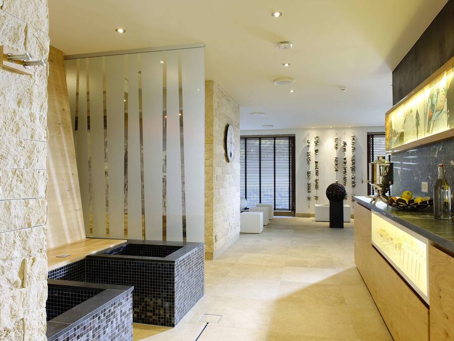 Una cocina moderna con encimera de madera, salpicadero de azulejos negros y pared de piedra. Hay una partición de vidrio con líneas verticales y un reloj en la pared.
