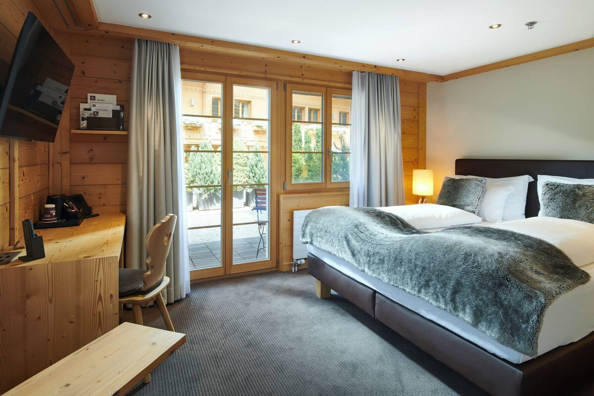 Una habitació d'hotel amb interior de fusta, un llit amb una manta de pell de color gris, un escriptori amb una cadira, un televisor fixat a la paret i una finestra amb cortines.