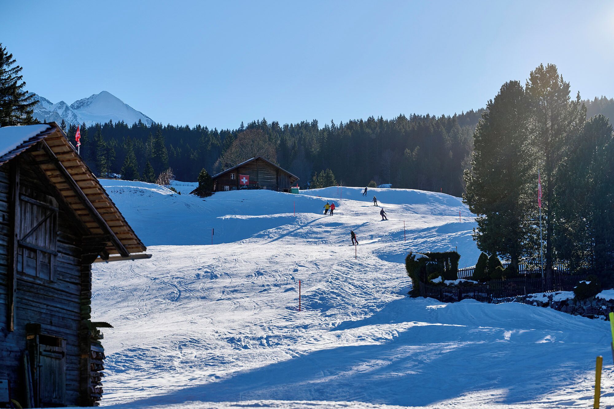 Eine Holzhütte mit Schweizer Flagge, einer Skipiste und einem Berg im Hintergrund.