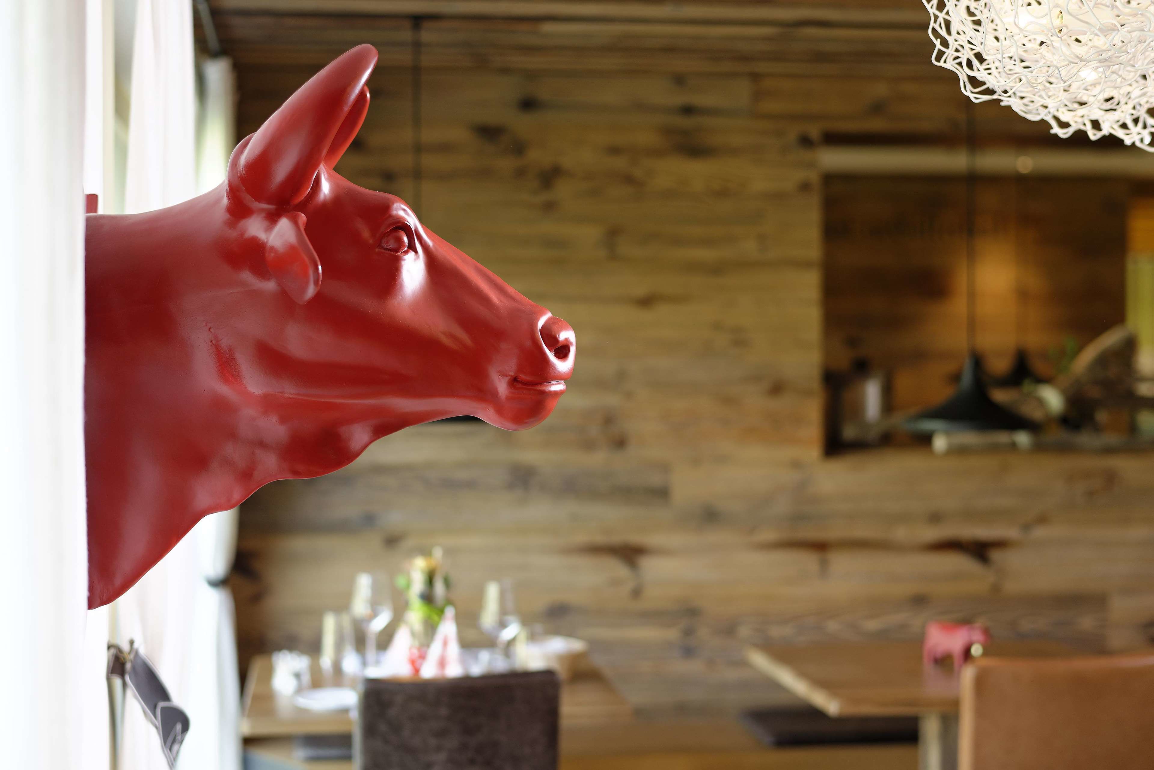 A red cow head sculpture mounted on a wall in a rustic interior with wooden paneling and a dining area in the background.