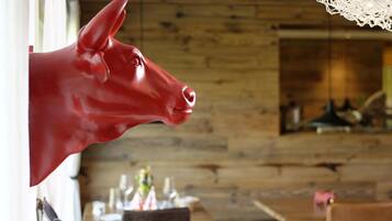 A red cow head sculpture mounted on a wall in a rustic interior with wooden paneling and a dining area in the background.