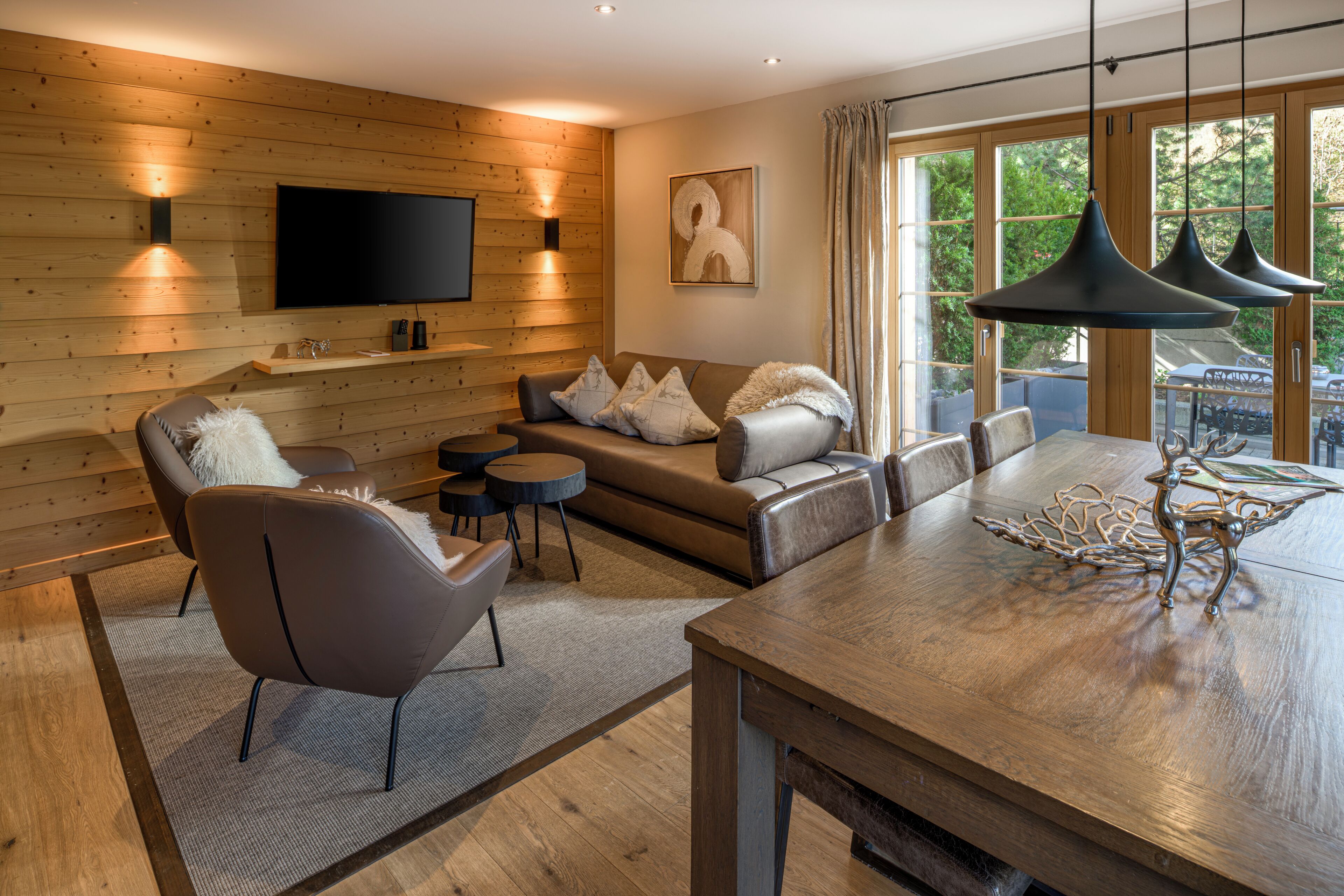A modern living room with a wooden wall, a flat-screen TV, a sofa set, a dining table with a metal sculpture, and a large window with curtains.