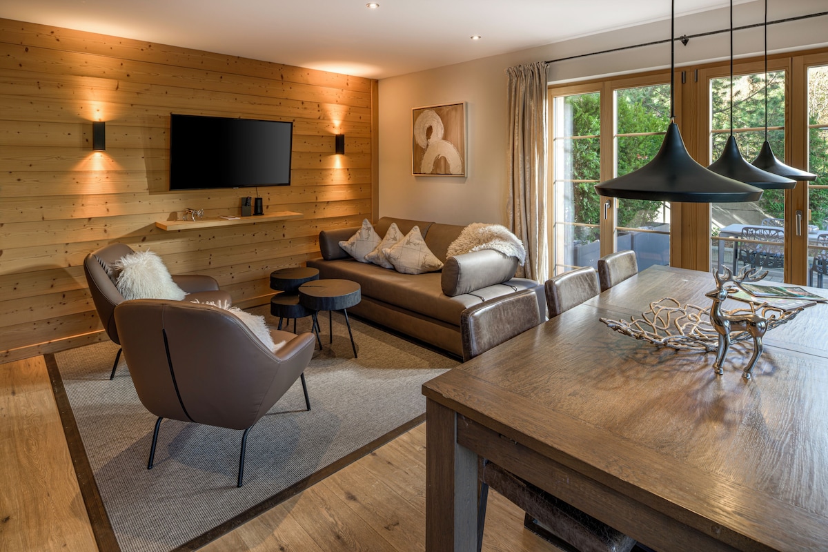 Una sala moderna con pared de madera, televisor de pantalla plana, conjunto de sofá, mesa de comedor y una ventana grande con cortinas.
