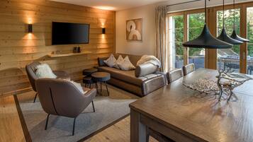 A modern living room with a wooden wall, a flat-screen TV, a sofa set, a dining table with a metal sculpture, and a large window with curtains.